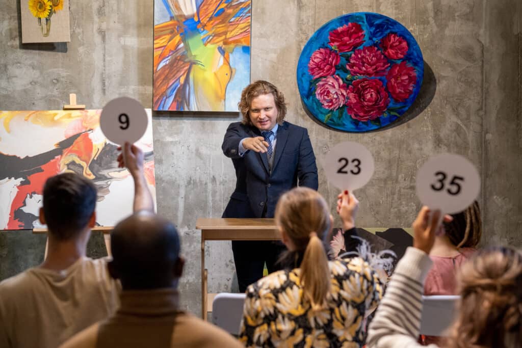 Mature male auctioneer in formal wear standing by tribune and pointing at one of people with auction paddles at a fundraising event