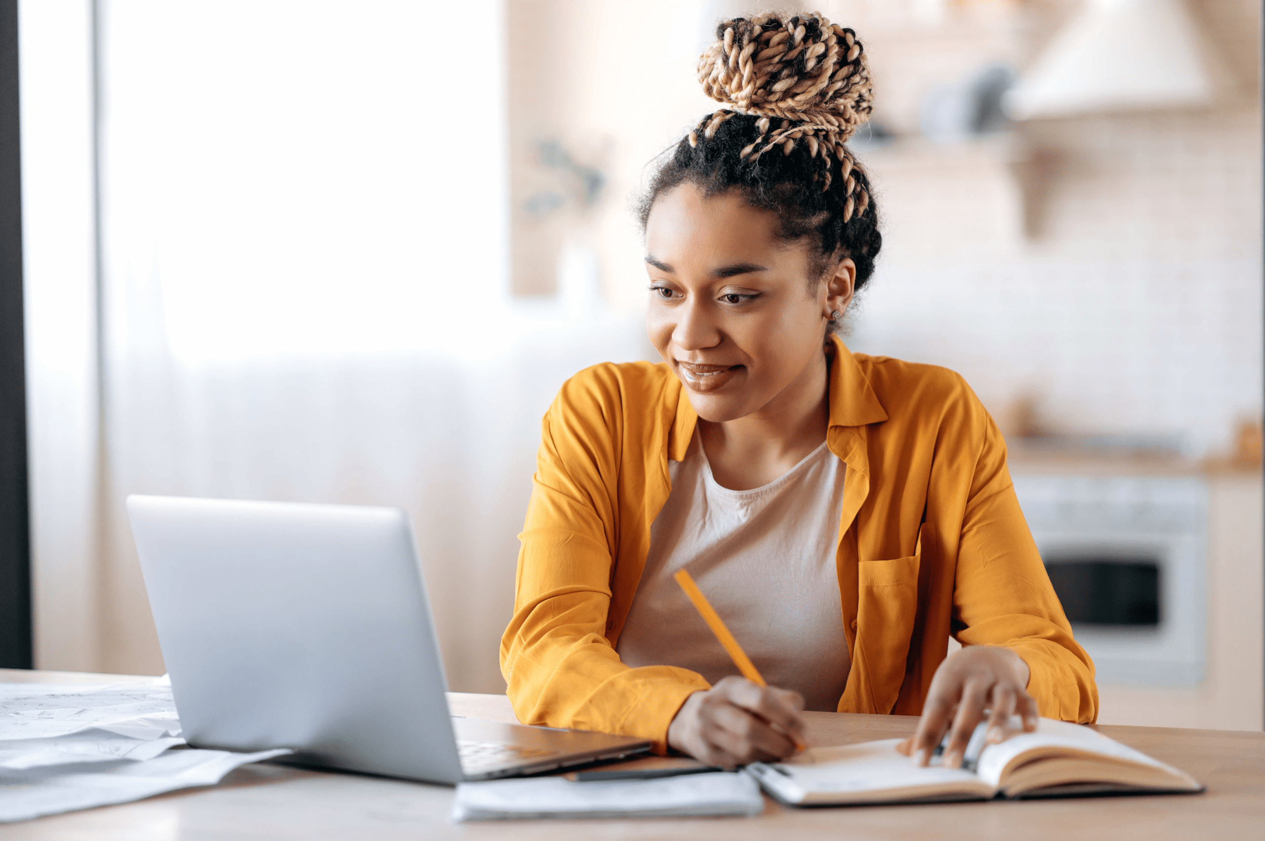 girl with a bun taking notes while watching a class on her laptop