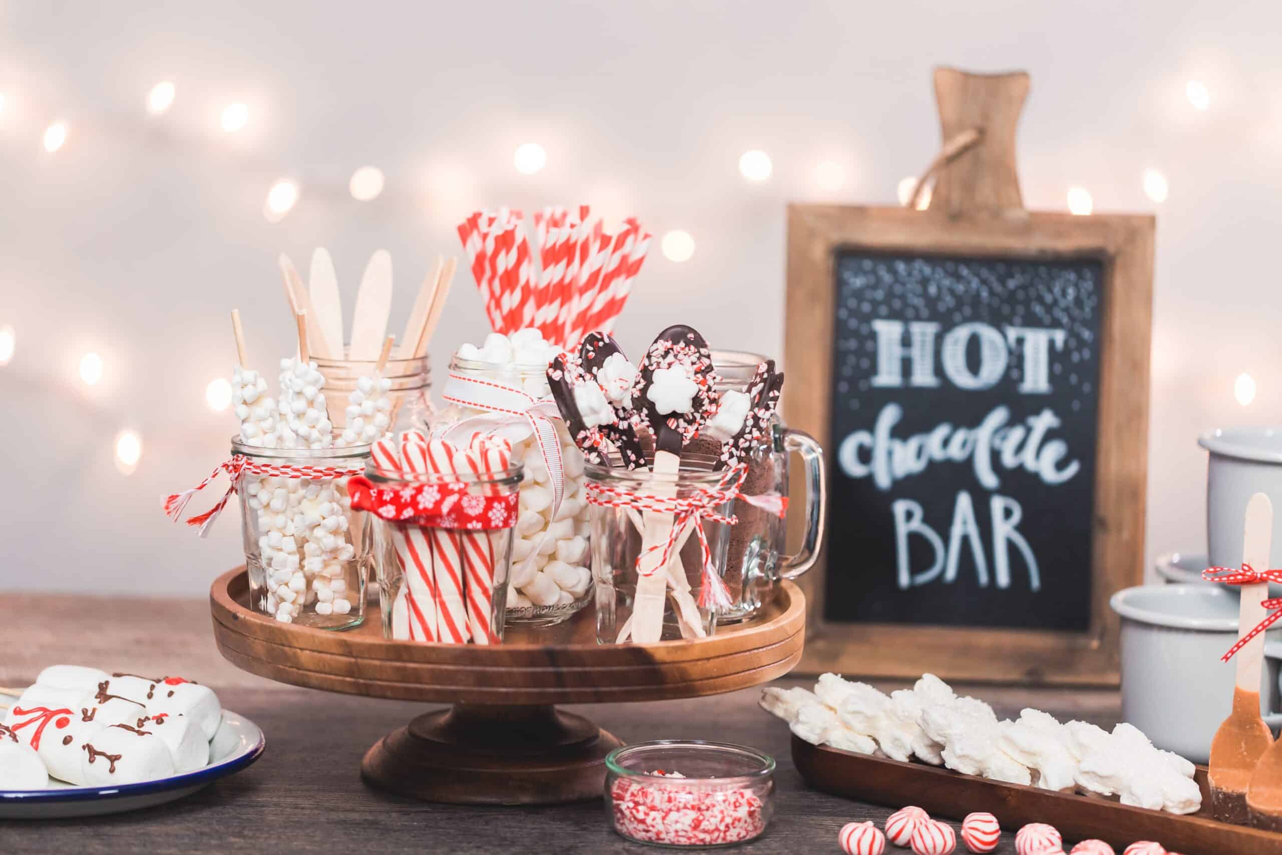 A table with hot cocoa toppings such as chocolate stirrers, peppermint sticks, and marshmallows with a sign that says "Hot Chocolate Bar"